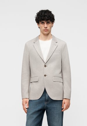 Fiatal férfi göndör hajjal, világos bézs kétsoros zakóban, fehér ingben és kék farmerben, egyszerű háttér előtt.