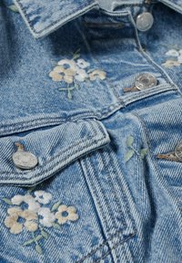 Chaqueta de mezclilla en azul claro con bordados florales en beige, blanco y verde. Presenta herrajes plateados y un cuello estructurado.