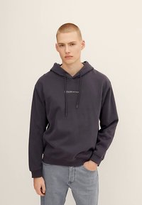 Tmavomodrá sivá mikina s kapucňou so šnúrkami, klokaním vreckom a vyšitým logom na hrudi. Kombinovaná so svetlosivými džínsami.