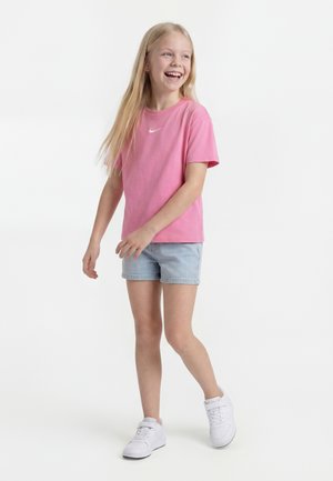 Lachend meisje met lang blond haar, een roze Nike T-shirt, lichtblauwe spijkershorts en witte sneakers, staand op een witte achtergrond.