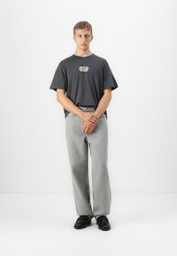 Young man standing with hands clasped, wearing a black "Wild Soul" t-shirt, light gray jeans, and black shoes against a plain white background.
