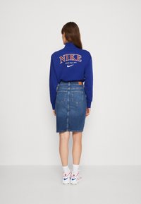 Blå sweatshirt med "NIKE"-logotyp, kombinerad med en figurnära jeanskjol. Vita sneakers med röda och blå detaljer, synlig sömnad och fickor.