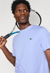 Ανοιχτό μοβ κοντομάνικο πουκάμισο από μαλακό ύφασμα, με μικρό πράσινο λογότυπο Lacoste και διακριτικό γραφικό σχέδιο στη μία πλευρά.