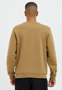 Brun sweatshirt med rund hals og ribbede manchetter og kant, der har en glat tekstur og et tags-Detalje på siden, vist fra bagsiden.