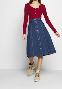 Top rojo de manga larga con detalle de botones, combinado con una falda vaquera hasta la rodilla con una fila de botones en la parte delantera. Zapatillas beige.