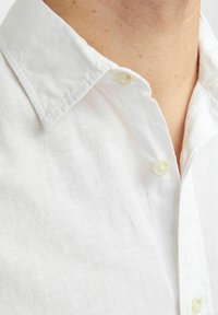 Camicia bianca di lino con colletto a punta, dotata di chiusura a bottoni e un tessuto a trama texturizzata. Il tessuto è leggero e traspirante.