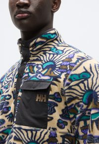 Φλις πούλοβερ σε κρεμ χρώμα με πολύχρωμα floral σχέδια. Μαύρη ταινία με κουμπιά και τσέπη στο στήθος με χρυσό λογότυπο «HH». Γιακαδάκι με έμφαση.