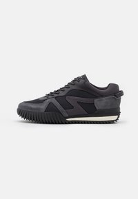 Chaussure de sport noire et grise avec un mélange de matériaux synthétiques lisses et de daim, dotée d'une semelle en caoutchouc dentelée et d'un design à lacets.