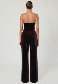 Burgundy bársony overál, pánt nélküli dizájnnal, testhez simuló fazonnal, széles lábakkal és hátsó cipzáros záródással. Két hátsó zseb található rajta.