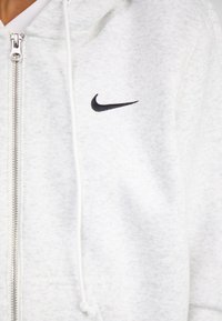 Grå hettegenser med glidelås laget av myk stoff med teksturert overflate, med en svart Nike-logo på brystet og justerbare hvite snorer.
