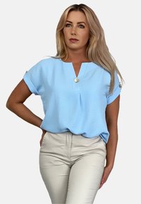 Jasnoniebieska krótkozasięgowa bluzka z dekoltem w serek i dekoracyjnymi złotymi guzikami. Noszona z dopasowanymi beżowymi spodniami. Gładka tekstura.