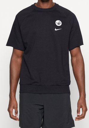 Homme portant un sweat-shirt noir à manches courtes avec un petit patch rond et un logo Nike blanc, associé à un short noir avec un logo Nike discret.