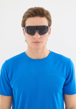 Nuori mies vaaleanruskeilla hiuksilla, päällään ylisuuret tummat urheilulasit ja kirkkaan sininen lyhythihainen urheilupaita, katsoo suoraan eteenpäin.