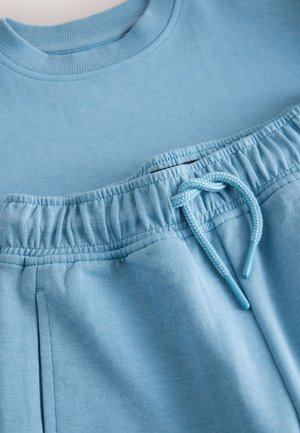 Világoskék pulóver anyag, kerek nyakkivágással, rugalmas derékpánttal és állítható húzózsinórral, amelynek végei kékek, két oldalzsebbel.