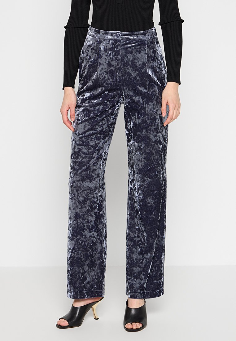 Vrouw in gekreukt blauw fluweel met rechte pijpen, een zwarte geribbelde lange mouwen top en zwarte open teen muilen met gouden stiletto-hakken.