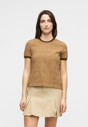 Femeie cu păr lung și închis la culoare, purtând un top maro cu mâneci scurte și model, cu margini închise la culoare, și o fustă plisată bej, în picioare pe un fundal simplu.