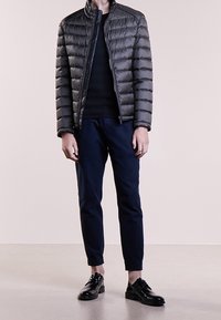 Veste rembourrée grise avec coutures horizontales, pull noir, pantalon bleu marine et chaussures en cuir noir. Le modèle pose devant un fond neutre.