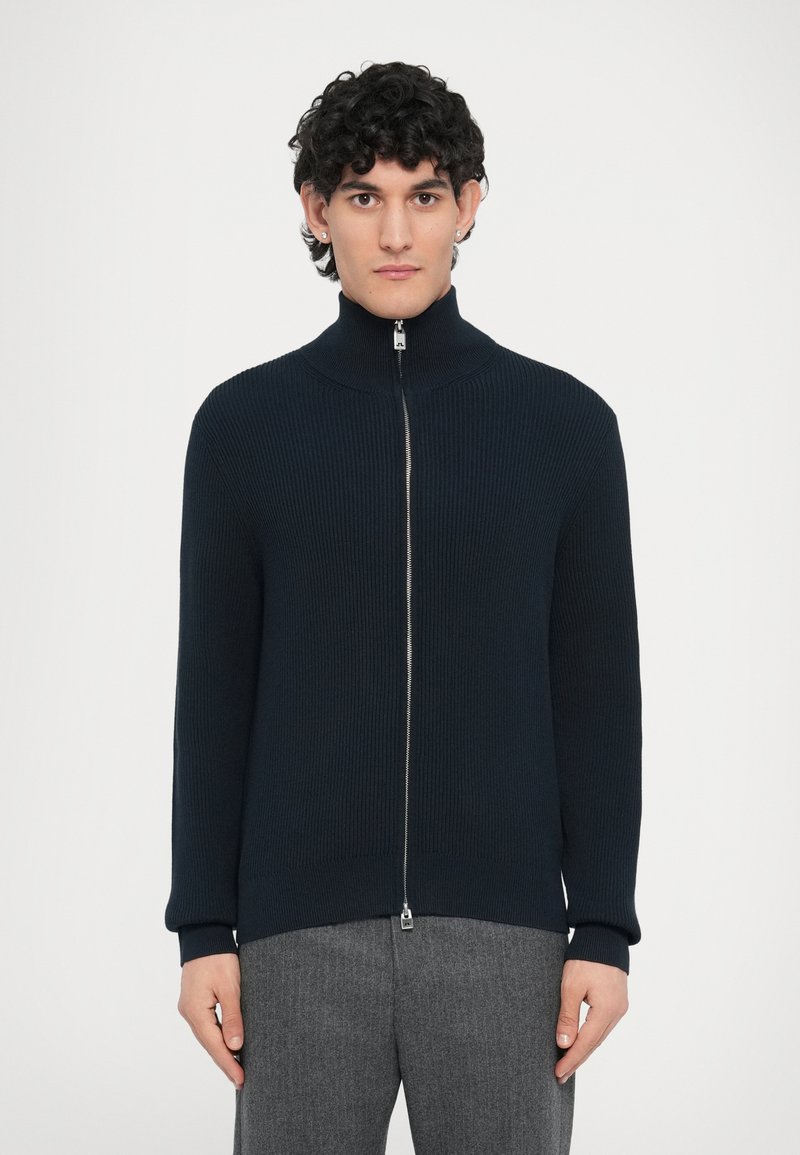 Μπλε ναυτικό πλεκτό πουλόβερ με ριμπ ύφασμα, ψηλό γιακά, ολόκληρο φερμουάρ και εφαρμοστά μανίκια, σε συνδυασμό με γκρι παντελόνια, εμφανίζεται σε λευκό φόντο.