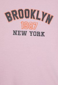 Roza pamučna majica s podebljanim crnim i narančastim natpisom "BROOKLYN 1987 NEW YORK." Glatka tekstura i opušten kroj.