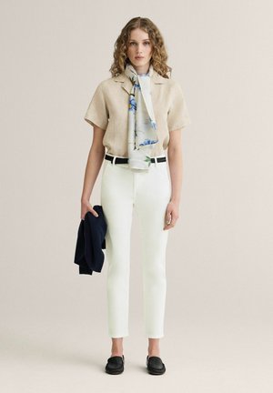 Kvinde med krøllet lyst hår iført beige kortærmet skjorte, hvide bukser med sort bælte, blomsteret tørklæde, sorte sko, holder en mørk jakke i venstre hånd.