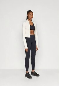 Kvinne som står, iført svart sport BH, svarte leggings, hvit kort jakke og svarte joggesko mot en ensfarget lys bakgrunn.