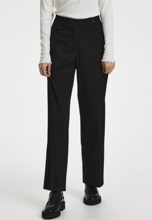 Kvinde iført sorte bukser med lige ben, hvid ribbet langærmet top og sorte chunky loafers, stående mod en ensfarvet baggrund.
