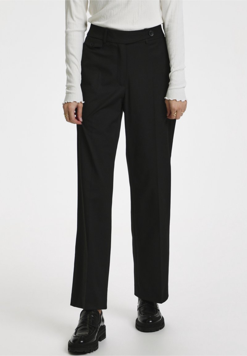 Femme portant un pantalon noir droit, un haut blanc à manches longues côtelé et des mocassins noirs épais, debout devant un fond uni.