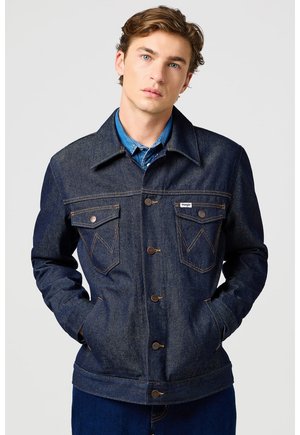 Młody mężczyzna o jasnobrązowych włosach, ubrany w ciemną kurtkę dżinsową na niebieską koszulę, ręce w kieszeniach, neutralne tło.