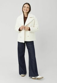 Caban en laine blanc croisé avec grands revers, porté sur un pull marron, assorti à un pantalon en denim foncé à jambes larges et des baskets blanches.