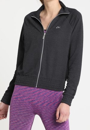 Schwarze sportliche Jacke mit Reißverschluss aus strukturiertem Stoff und geripptem Saum, getragen über lila gemusterten Leggings, mit Stehkragen.
