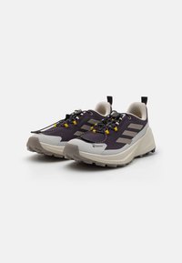 Chaussures de trail Gore-Tex en violet foncé et gris, avec une tige texturée, une semelle en caoutchouc avec des crampons agressifs, et des accents jaunes sur les lacets.
