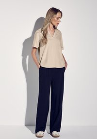 Beige Kurzarmoberteil mit V-Ausschnitt, kombiniert mit marineblauen weit geschnittenen Hosen und weißen Sneakers. Weicher Stoff mit lockerer Passform.