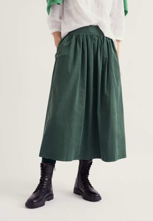 Personne portant un chemisier blanc, une longue jupe verte et des bottines noires à lacets, debout les mains dans les poches devant un fond uni.