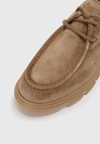 Bateau en daim brun avec un bout arrondi, des coutures ton sur ton et une épaisse semelle en caoutchouc beige. Les lacets sont noués sur le dessus.