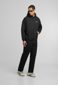 Sudadera negra con bolsillo frontal y logotipo, combinada con pantalones de chándal negros a juego. Presenta zapatillas con acentos en blanco y verde.