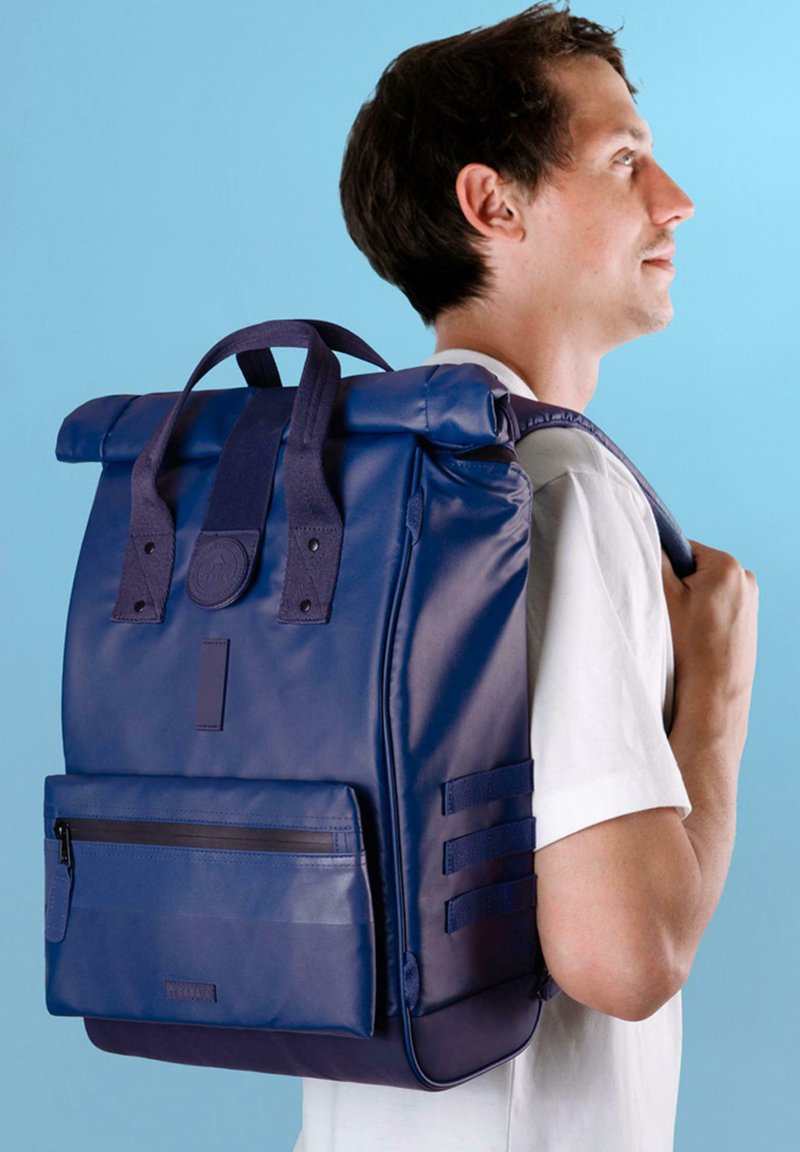Marinblå rulltopprygsekk med slitesterk stoff, to bærehåndtak, sidehåndtak og frontlomme med glidelås, plassert mot en lys blå bakgrunn.