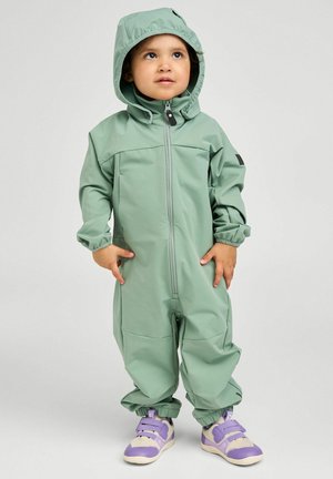 Kleinkind trägt hellgrünen Kapuzenregenanzug und lila Turnschuhe, steht mit Händen in der Nähe der Taschen und blickt leicht nach oben.