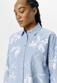 Camicia a righe blu e bianche con motivi floreali bianchi; vestibilità ampia, design con bottoni e colletto classico. Tessuto morbido.