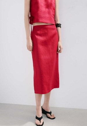 Mujer con top sin mangas rojo y falda midi a juego, accesorios con pulseras negras y sandalias de cuña negras, posando contra un fondo liso.