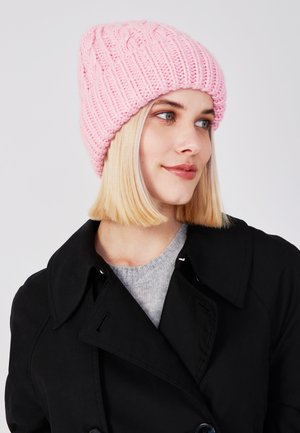 Gebreide roze beanie met een geribbelde textuur en kabelpatroon. Draagt een zwarte jas en een grijze trui, wat een casual winterlook laat zien.