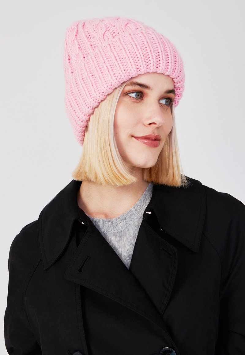 Gebreide roze beanie met een geribbelde textuur en kabelpatroon. Draagt een zwarte jas en een grijze trui, wat een casual winterlook laat zien.