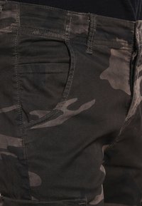 Zbliżenie na spodenki w ciemnoszarym i czarnym kamuflażu, pokazujące przednią kieszeń i szlufki na pasek, noszone przez osobę w czarnej górze.