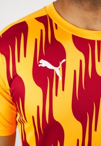 Primo piano di una persona che indossa una maglietta sportiva Puma con un motivo a fiamma gialla e rossa, con il logo Puma bianco sul petto.