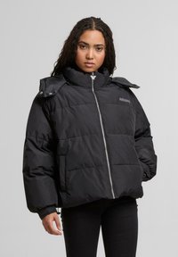 Giovane donna che indossa un piumino nero con zip e colletto alto, in piedi con una mano in tasca contro uno sfondo grigio semplice.