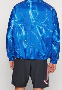 Osoba w niebieskiej kurtce z białymi wzorami w spiralę oraz czarnych szortach z różowym logo Nike, zwrócona plecami do aparatu.