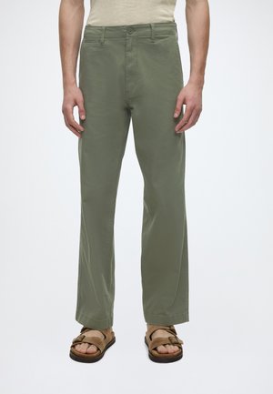 Homme portant un pantalon droit vert olive et des sandales beiges, debout devant un fond blanc uni.