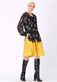 Blouse noire transparente avec motif floral, col volanté et boutons, associée à une jupe jaune vif mi-longue et des bottes noires jusqu'aux genoux.