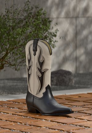 Stivale cowboy alto bicolore con punta nera appuntita e ricamo complesso a fiamma beige e nero, posato su terreno esterno di mattoni e ghiaia.