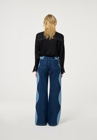 Donker shirt in de taille gestopt in breedvallende blauwe denim jeans met contrasterende lichtblauwe golfpatronen langs de zijkanten. Schone, minimalistische uitstraling.