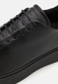 Black leather shoe with flat laces, rounded toe, and minimalistic design. Features a textured surface and subtle branding on the upper.